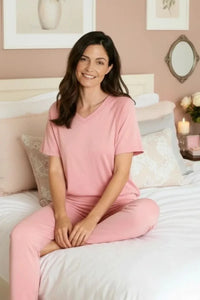 Woman in pink short sleeve loungewear set sitting cross-legged on white bed in cozy bedroom